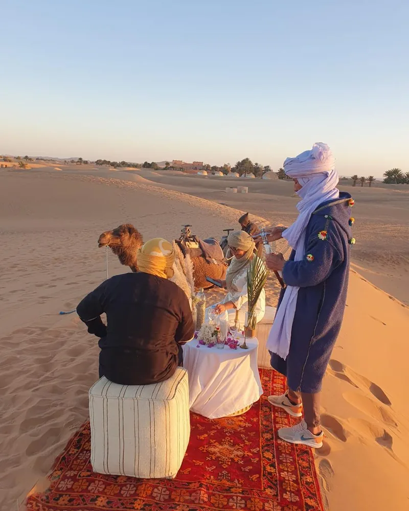 Un guía bereber con turbante atiende a dos viajeros sobre una alfombra en las dunas del Sahara