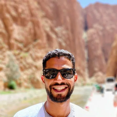 Ibrahim, fundador y guía de Morocco Soul Travel, sonríe con gafas de sol ante las paredes rojas de un desfiladero